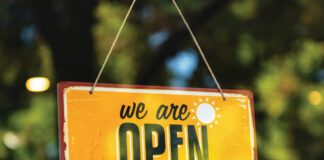 A yellow sign reads “we are OPEN,” and hangs on twine in front of a window.