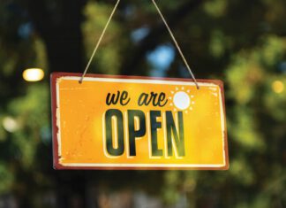 A yellow sign reads “we are OPEN,” and hangs on twine in front of a window.