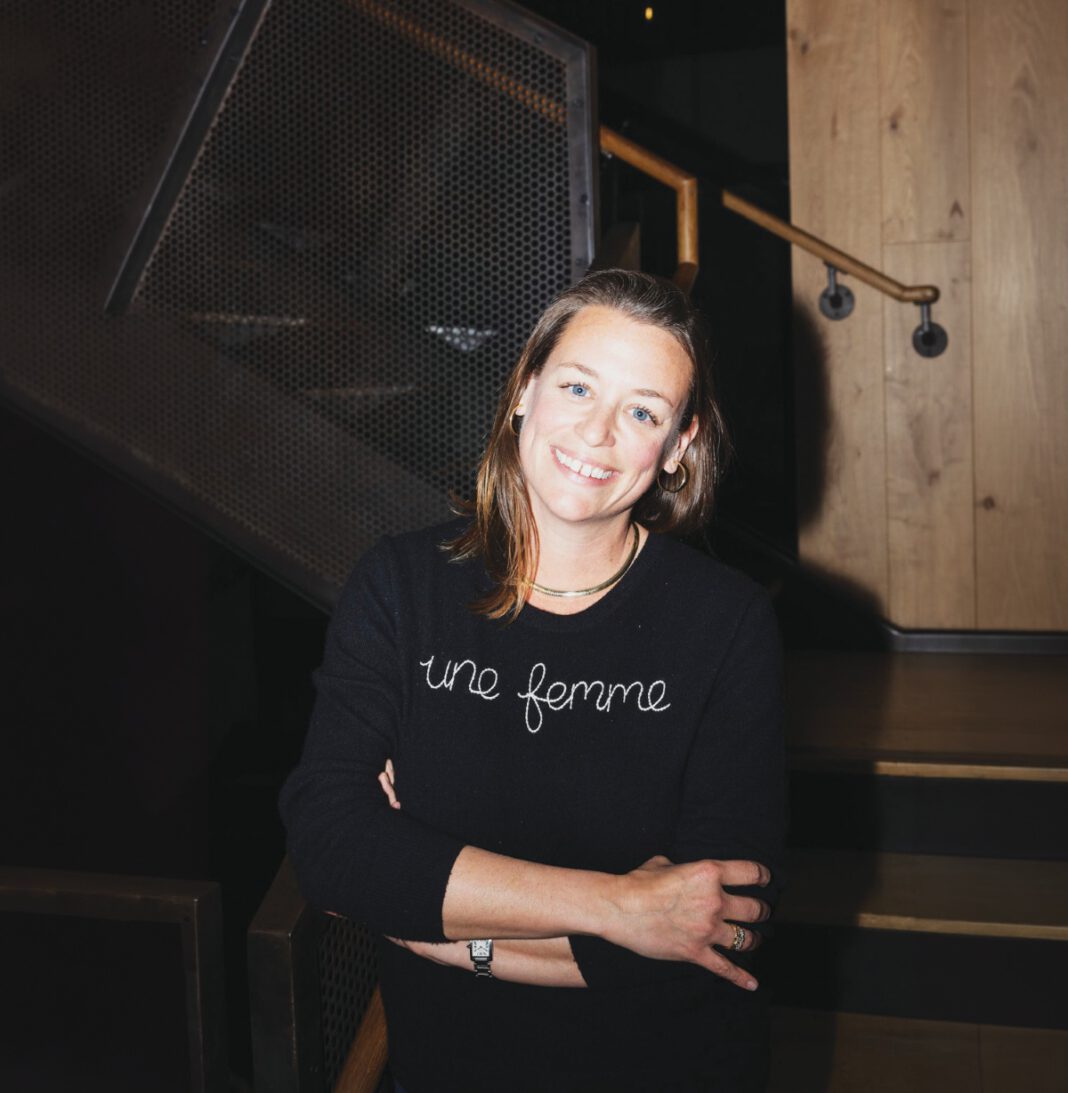 A smiling woman with shoulder-length hair wearing a black shirt that reads “une femme” stands in front of a wood staircase.