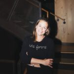 Pop the Can: Jen Pelka Sparkles with Wine Business A smiling woman with shoulder-length hair wearing a black shirt that reads “une femme” stands in front of a wood staircase.