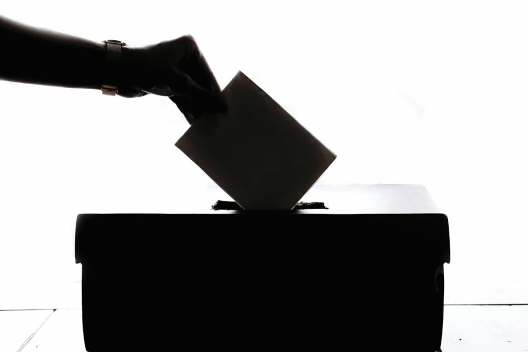 In silhouette, a hand drops a folded ballot into a black box.