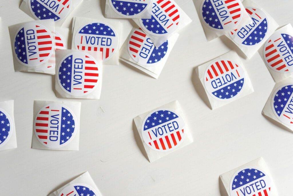 Round red, white and blue “I Voted” stickers are scattered on a white table.