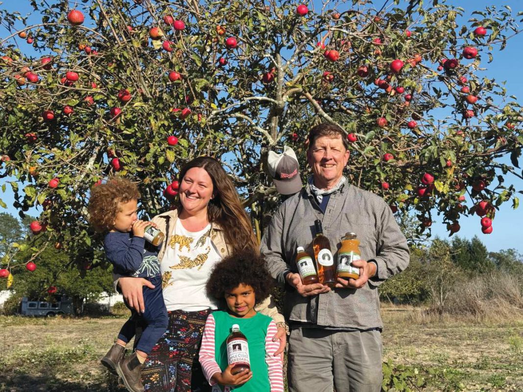 A smiling woman holding a toddler, a girl holding a bottle and a smiling man holding bottles stand in front of an apple tree full of red apples.