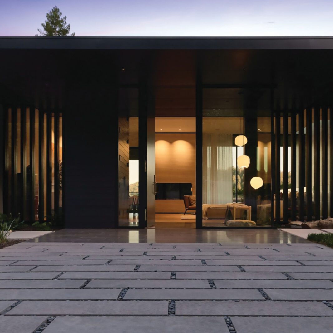 A stone-paved walkway leads to single-story dark brown modern house with floor-to-ceiling glass windows.