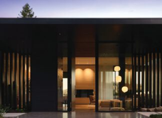 A stone-paved walkway leads to single-story dark brown modern house with floor-to-ceiling glass windows.