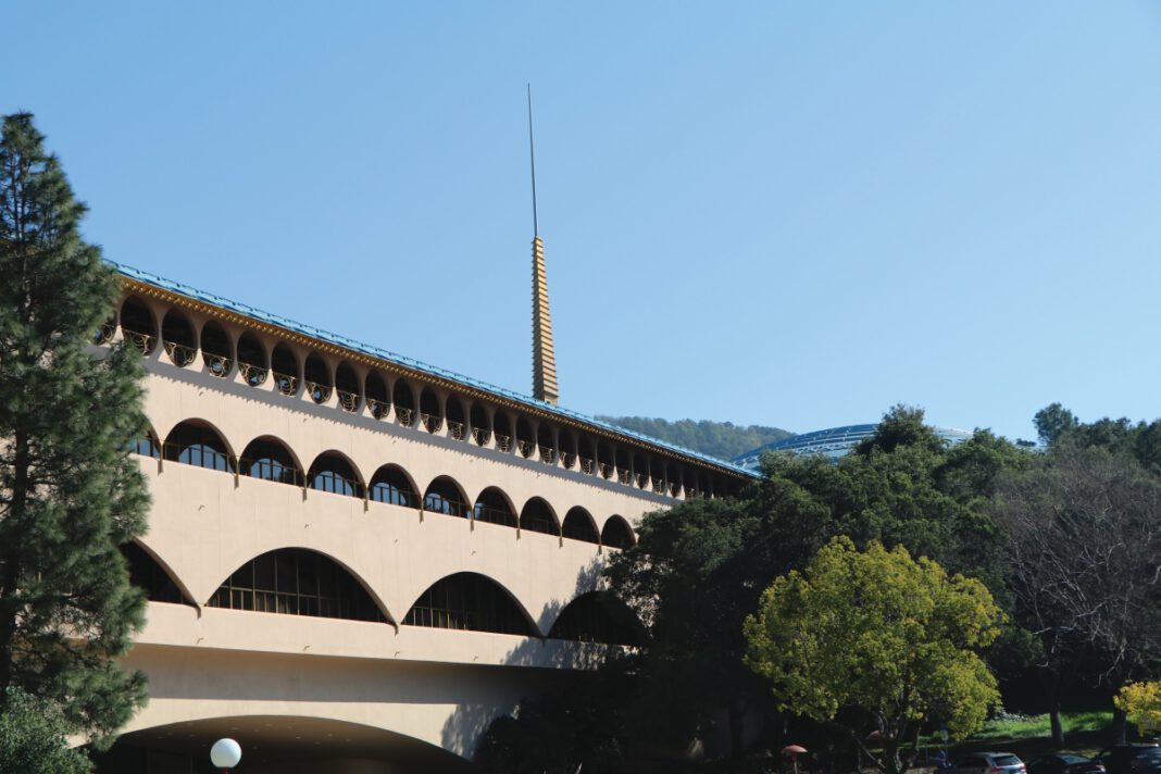A long cream-colored building with rows of arched windows and a tall gold spire with blue sky above it.
