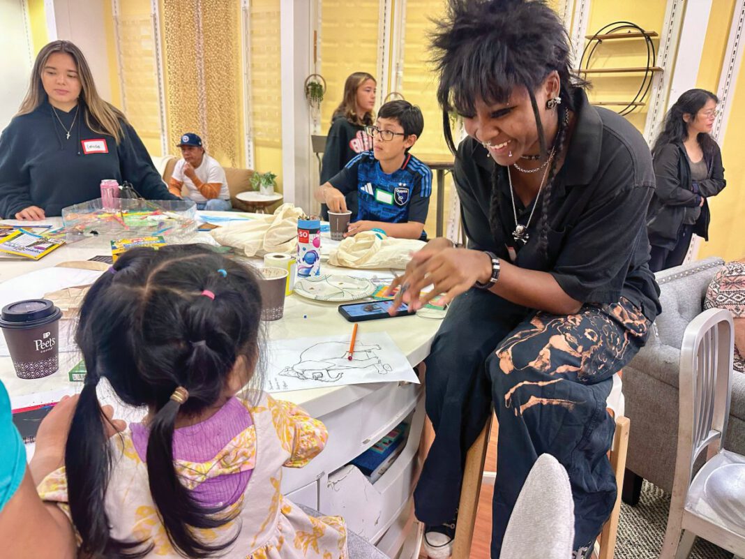 A young woman with spiky black hair, braids and piercings sitting at a table, laughing with a little girl seen from behind.