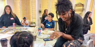 A young woman with spiky black hair, braids and piercings sitting at a table, laughing with a little girl seen from behind.