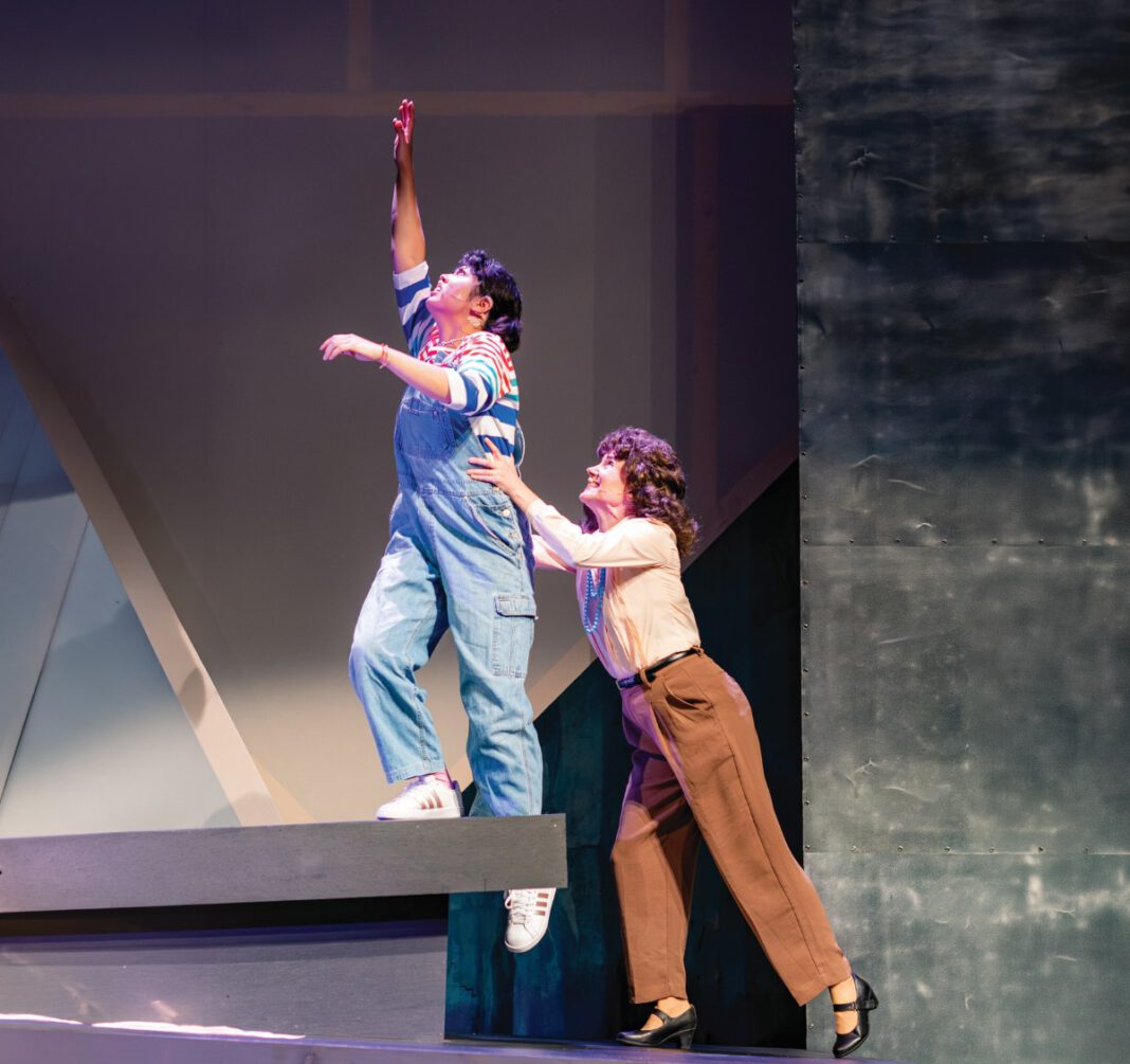 One female actor, standing on a gray ledge, is held up by another female performer.