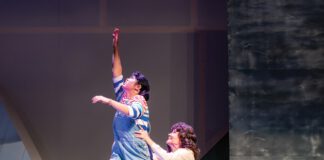 One female actor, standing on a gray ledge, is held up by another female performer.