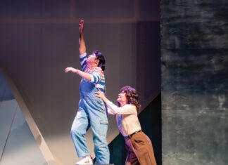 One female actor, standing on a gray ledge, is held up by another female performer.