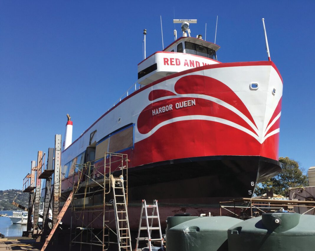 A red and white boat named Harbor Queen sits out of the water in a boatyard, surrounded by scaffolding and ladders.