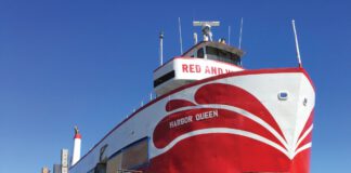 A red and white boat named Harbor Queen sits out of the water in a boatyard, surrounded by scaffolding and ladders.