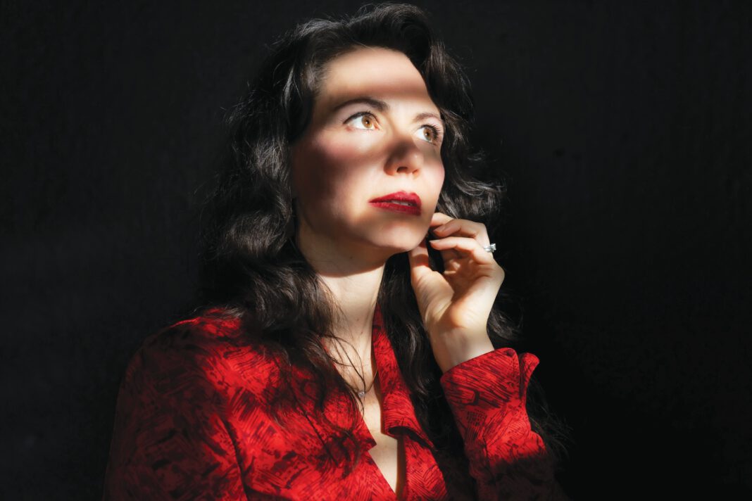 A woman with long dark hair and red lipstick, wearing a red blouse, has one hand on her chin.