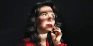A woman with long dark hair and red lipstick, wearing a red blouse, has one hand on her chin.