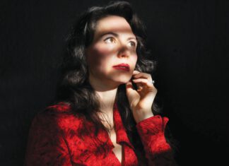 A woman with long dark hair and red lipstick, wearing a red blouse, has one hand on her chin.