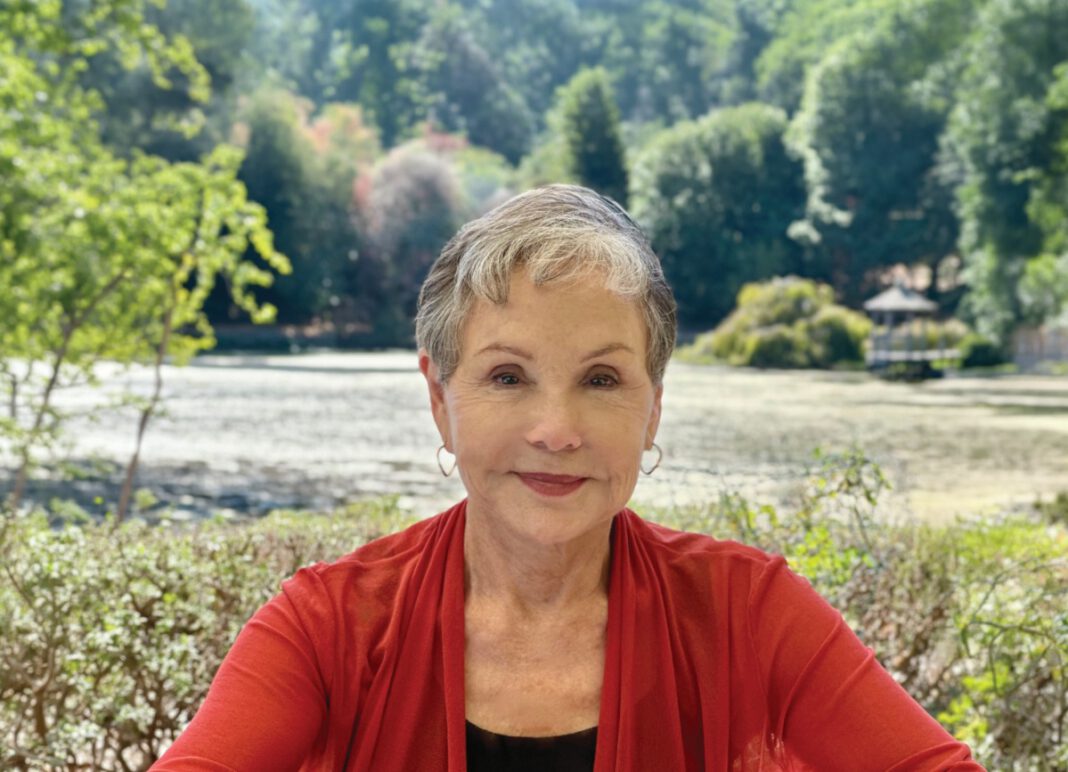 A smiling woman with short gray hair, hoop earrings and a red cardigan sits outside in front of trees.