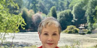 A smiling woman with short gray hair, hoop earrings and a red cardigan sits outside in front of trees.