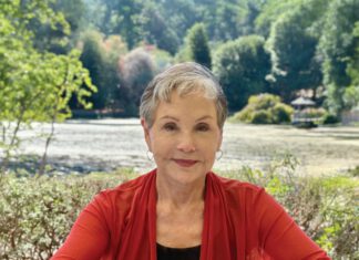 Dream State: Artist, Writer, Poet Ayris Hatton A smiling woman with short gray hair, hoop earrings and a red cardigan sits outside in front of trees.