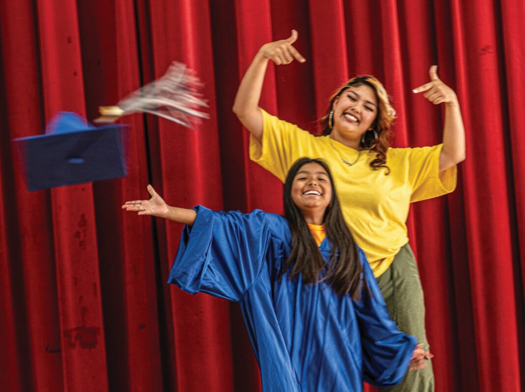 A smiling girl wearing a blue graduation gown throws her cap into the air while a smiling woman in a yellow shirt stands behind her gesturing.