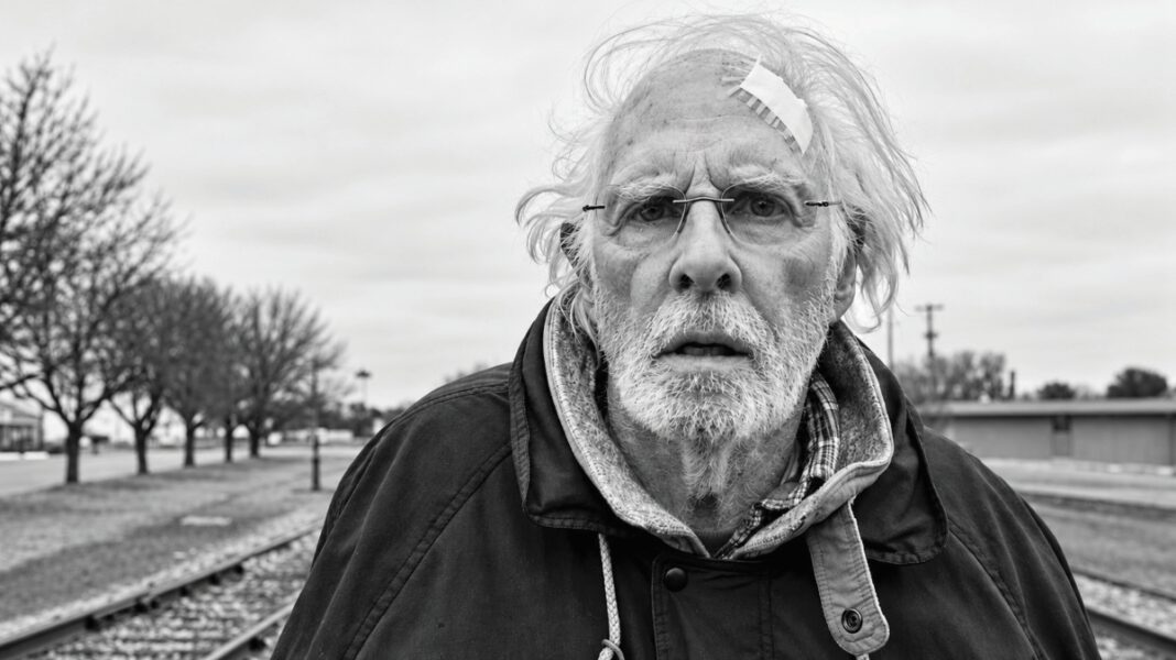 A black-and-white photo of an older man with white hair, a white beard, eyeglasses and a bandage on his forehead standing near railroad tracks.