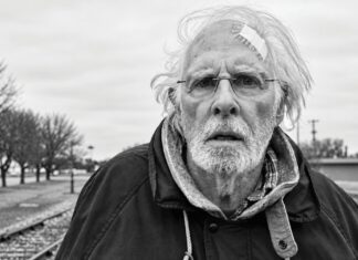 A black-and-white photo of an older man with white hair, a white beard, eyeglasses and a bandage on his forehead standing near railroad tracks.