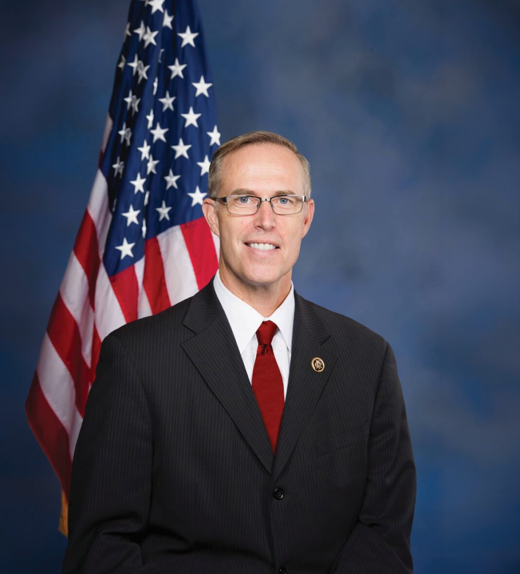A smiling man with short gray hair and glasses wears a dark suit, white shirt and red tie and standing in front of an American flag in front of a blue background.