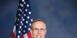 A smiling man with short gray hair and glasses wears a dark suit, white shirt and red tie and standing in front of an American flag in front of a blue background.