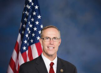 Rep. Jared Huffman Connects Household Costs, Congress and Christian Nationalism A smiling man with short gray hair and glasses wears a dark suit, white shirt and red tie and standing in front of an American flag in front of a blue background.