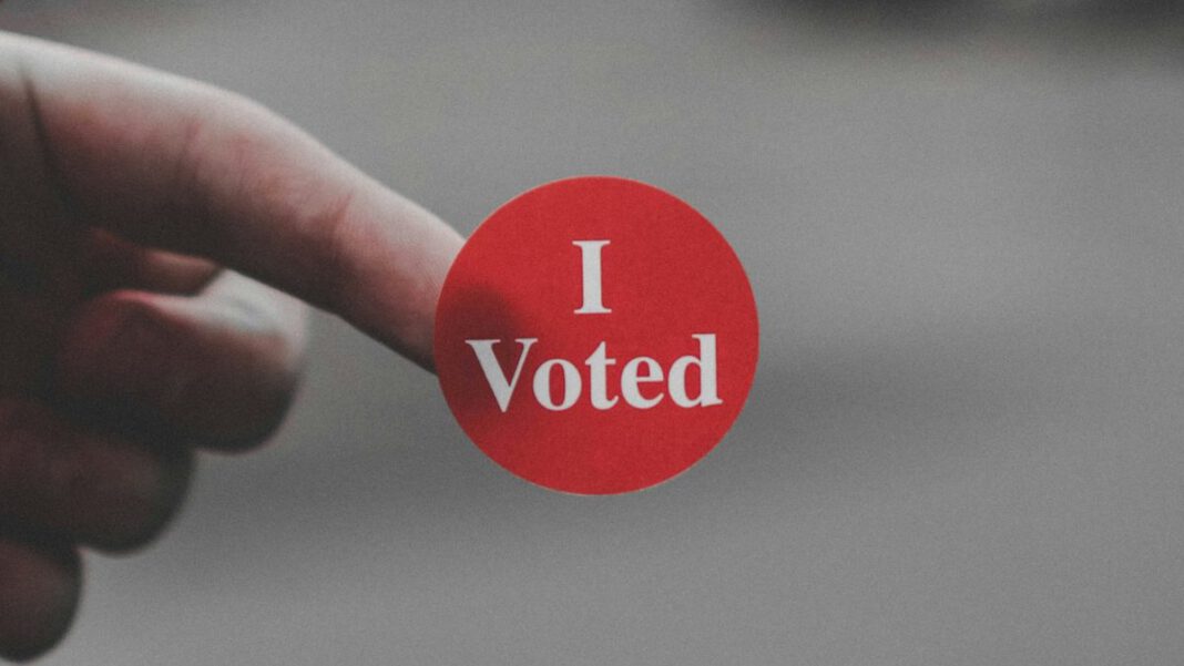 A hand holds a round red sticker that says “I Voted” in front of a blurry gray background.